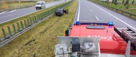 Zdjęcie zrobione z dachu samochodu pożarniczego. Na zdjęciu widać samochód osobowy poza drogą w pozycji na kołach, który stoi na pasie zieleni bezpośrednio przy barierach energochłonnych