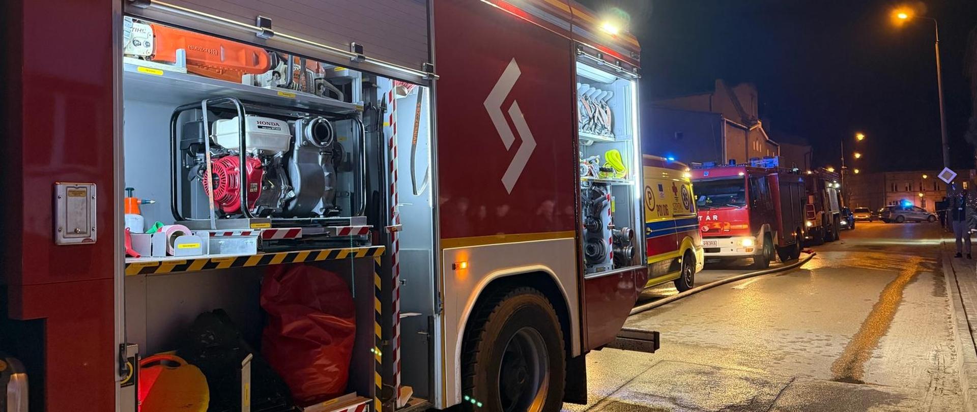Na zdjęciu widać trzy samochody pożarnicze, które zostały zadysponowane do zdarzenia. Stoją one na drodze przed budynkiem, pomiędzy nimi widać również karetkę pogotowia. Jest pora nocna, świecą latarnie uliczne.