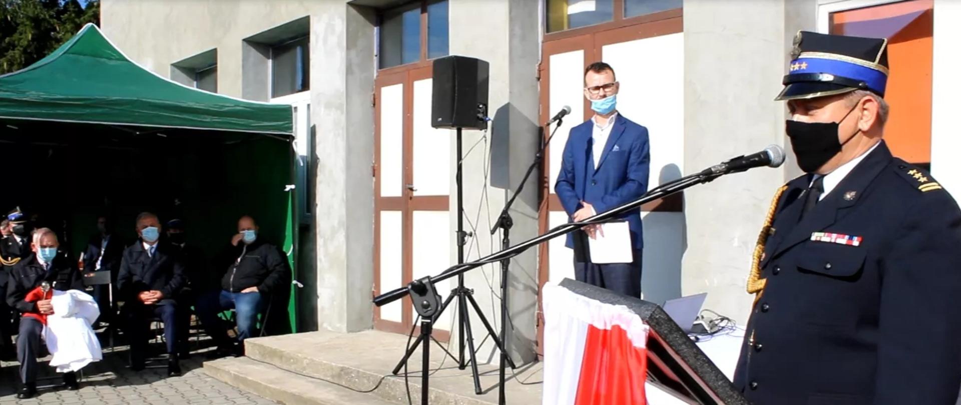 Na zewnątrz budynku z mównicy przemawia Komendant Wojewódzki PSP. na drugim planie pod namiotem plenerowym na ławkach siedzą zgromadzeni goście. 