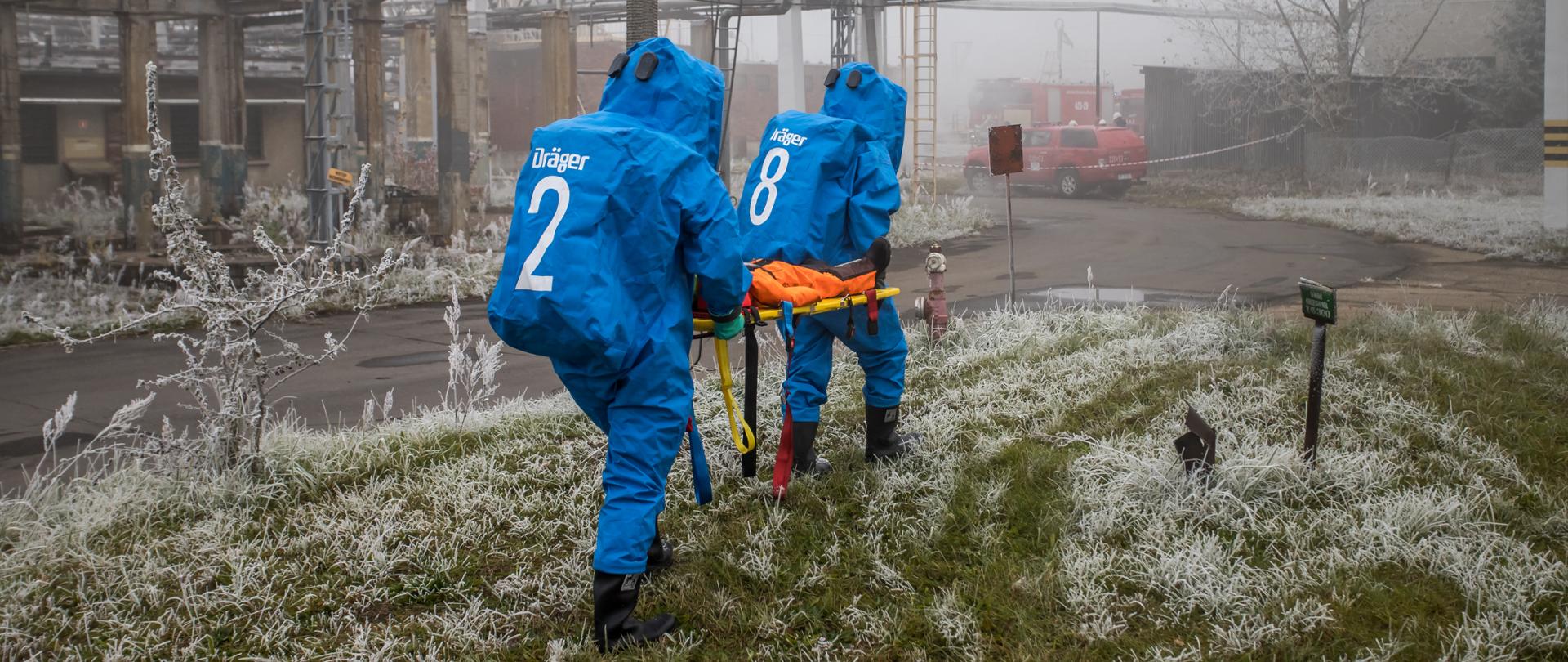 Zdjęcie przedstawia strażaków Specjalistycznej Grupy Chemiczno-Ekologicznej "Kędzierzyn-Koźle" w chemicznych ubraniach gazoszczelnych koloru niebieskiego (dwóch strażaków) idących i niosących manekina na desce ortopedycznej koloru żółtego. Na plecach strażaków napis koloru białego Drager i numery 2 u jednego z nich i 8 u drugiego strażaka. Strażacy idą po zroszonej trawie. W oddali widoczna droga asfaltowa. Zdjęcie wykonane na terenie jednego z zakładów. Nad strażakami widoczny rury rurociągów zakładu.