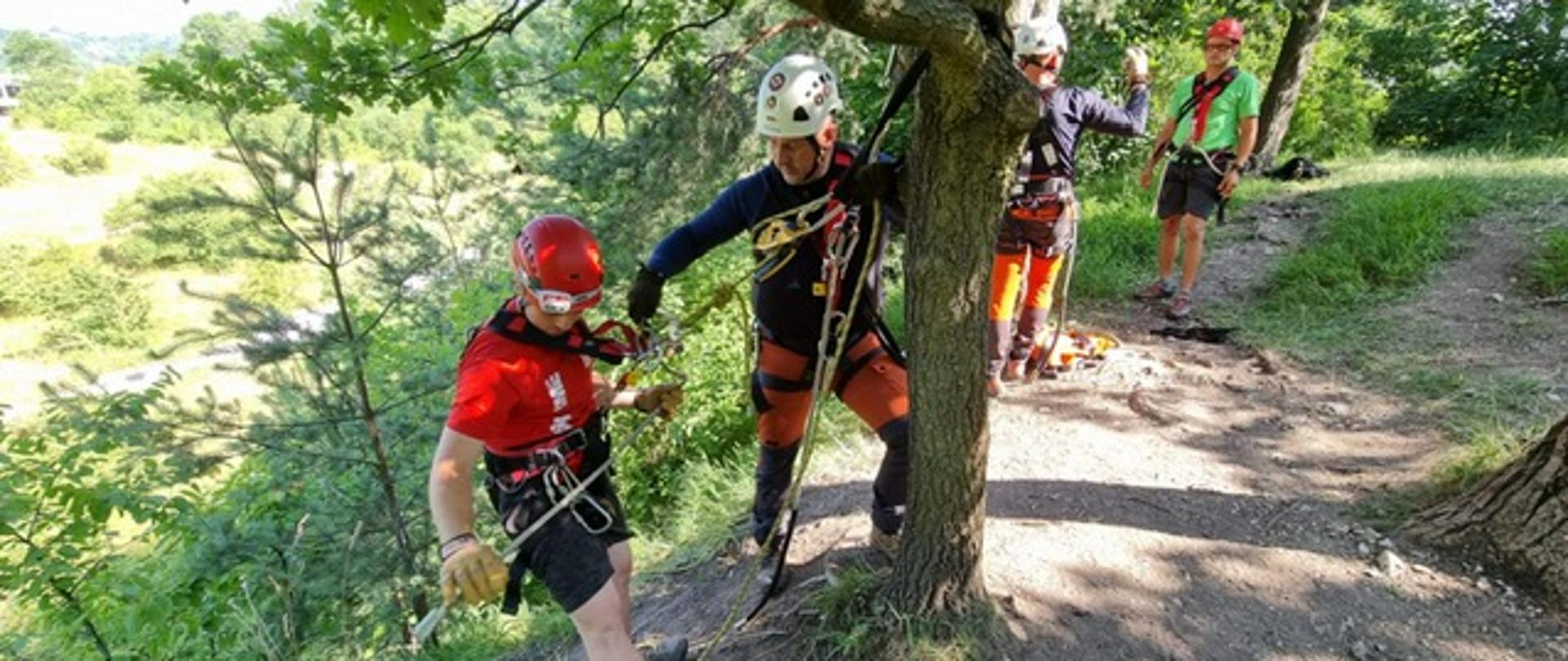 Mężczyźni ubrani w sprzęt do ratownictwa wysokościowego w asekuracji ćwiczą zejście po stromym ułożeniu terenu 