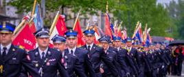 Strażacy Państwowej i Ochotniczej Straży Pożarnej w umundurowaniu galowym kroczą w szyku z licznymi pocztami sztandarowymi. W centrum widoczny jest strażak PSP w czapce z niebieskim otokiem i dystynkcjami oficerskimi, w tle – rzędy czerwonych sztandarów z hasłem „Bogu na chwałę, ludziom na ratunek”.
