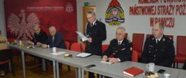 Zebranie sprawozdawcze OSP Rawicz. Świetlica Komendy Powiatowej PSP w Rawiczu. Za stołem prezydialnym siedzą zaproszeni goście oraz przedstawiciele zarządu miejscowej OSP. Przewodniczący zebrania na stojąco prowadzi obrady. Na stole leżą dokumenty.