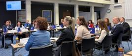 Uczestnicy programu podczas zajęć - kilkanaście osób siedzi przy stołach ustawionych w podkowę; w tle ekran multimedialny