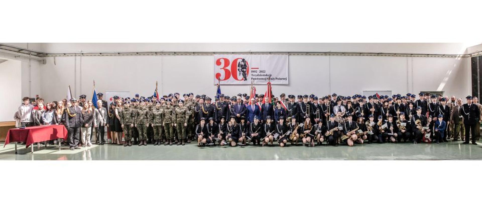 W pomieszczeniu stoi kilkaset osób. Z tyłu napis "30" i postać strażaka. Większość osób ubranych jest w mundury: strażackie, wojskowe oraz leśników.