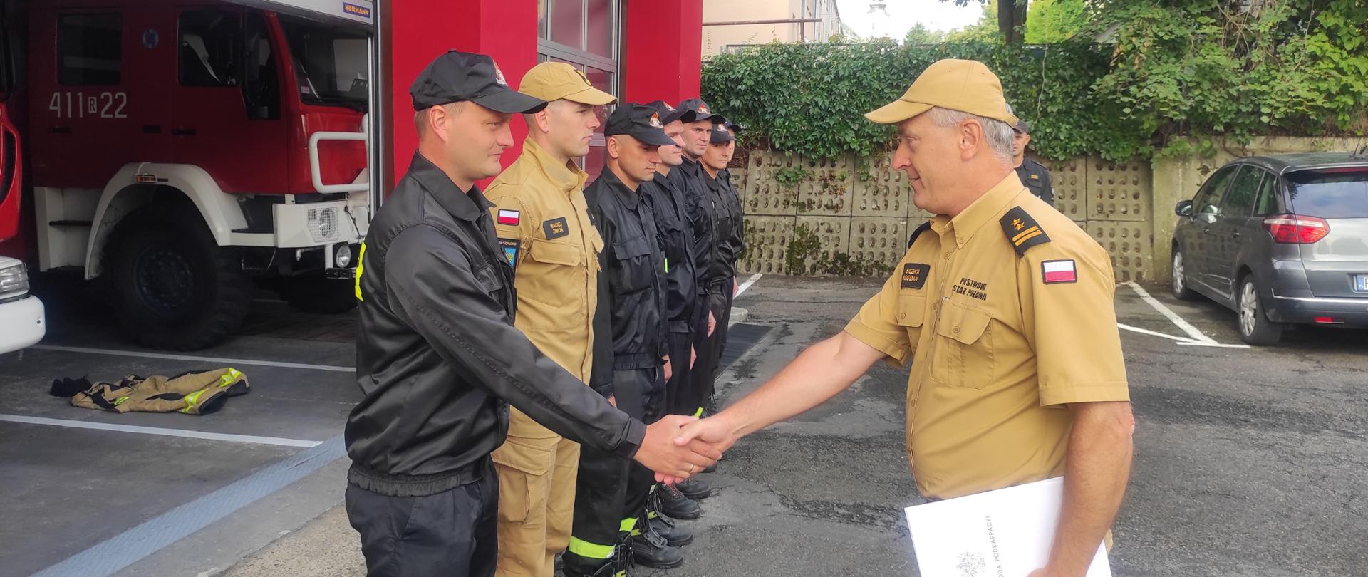 Kolorowa fotografia wykonana w pogodny dzień na zewnątrz. Przedstawia strażaków w ubraniach koszarowych i służbowych podczas zmiany służby. Na środku zastępca komendanta powiatowego który będzie wręcza pisemne podziękowania oraz dowódca przeprowadzający zmianę służby.