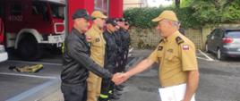Kolorowa fotografia wykonana w pogodny dzień na zewnątrz. Przedstawia strażaków w ubraniach koszarowych i służbowych podczas zmiany służby. Na środku zastępca komendanta powiatowego który będzie wręcza pisemne podziękowania oraz dowódca przeprowadzający zmianę służby.