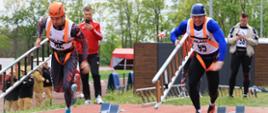 5. Dwóch zawodników startuje równolegle z drabinami hakowymi, biegnąc po torze w kierunku ściany ćwiczebnej. W tle widać innych uczestników i sędziów zawodów.
