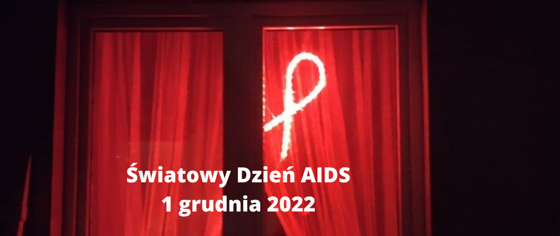 Zdjęcie przedstawia okno mieszkania w nocy, w oknie świeci się czerwona kokarda. Na zdjęciu znajduje się biały napis: Światowy Dzień AIDS 1 grudnia 2022. Happening