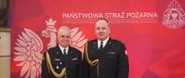 Dwóch funkcjonariuszy PSP w galowych mundurach stoi obok siebie na tle czerwonej planszy z godłem PSP. Obaj są wyprostowani, z rękami opuszczonymi wzdłuż ciała, patrzą w obiektyw aparatu. Mundury są starannie ułożone, z widocznymi dystynkcjami i odznaczeniami.