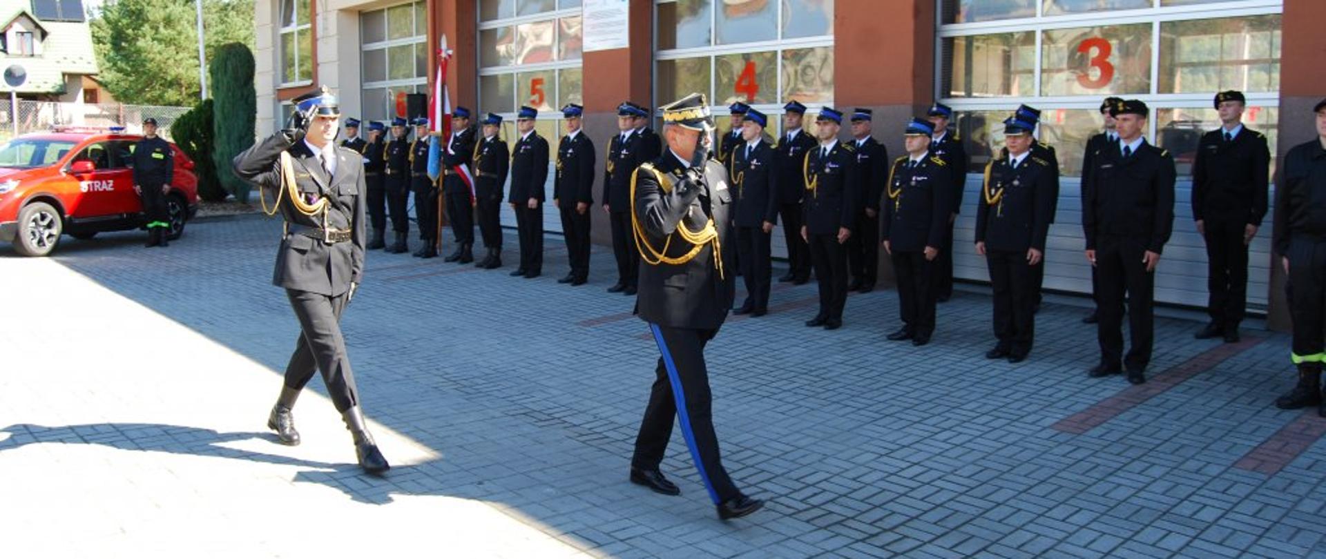 Zdjęcie przedstawia przegląd pododdziału. Nadbrygadier Andrzej Babiec Podkarpacki Komendant Wojewódzki Państwowej Straży Pożarnej w umundurowaniu galowym krokiem defiladowym oddając honor poprze salutowanie dokonuje przegląd pododdziału. Na zdjęciu w połowie pododdziału. Z nim po lewej stronie w odległości jednego kroku dowódca uroczystości w umundurowaniu galowym kapitan Marcin Bieszczad. Dowódca uroczystości oddaje honor poprzez salutowanie. W tle po lewej stronie nowy samochód operacyjny HONDA CRV. Obok samochodu w stoi strażak w umundurowaniu koszarowym. Dalej w prawo funkcjonariusze Komendy Powiatowej Państwowej Straży Pożarnej w Strzyżowie ustawieni w dwuszeregu. Z nimi widoczne bramy wyjazdowe garażów jednostki ratowniczo gaśniczej. Na początku pododdziału widoczne poczty flagowy i sztandarowy.