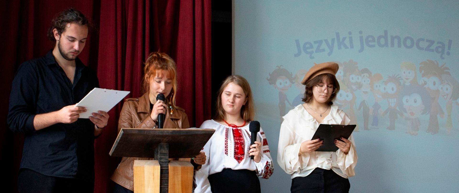 Czwórka młodych ludzi stoi na scenie przed czerwoną kurtyną i ekranem; trzymają w rękach mikrofony oraz notatniki, a kobieta w środku mówi do mikrofonu.
