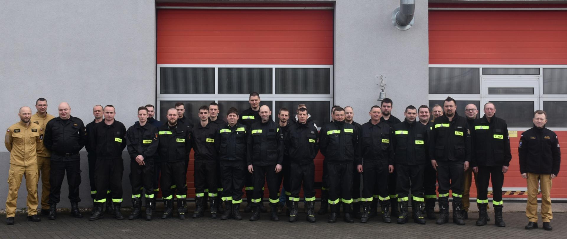 Zdjęcie wykonano w ciągu dnia na placu Komendy Powiatowej Państwowej Straży Pożarnej w Sępólnie Krajeńskim przed budynkiem garaży. Strażacy Ochotniczej Straży Pożarnej w ubraniach koszarowych wraz z Komendantami i Dowódcami ustawieni w dwuszeregu pozują do zdjęcia. 