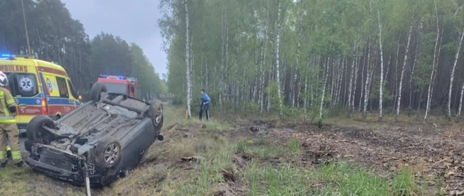 Uszkodzony pojazd po dachowaniu na poboczu, po lewej za nim karetka pogotowia