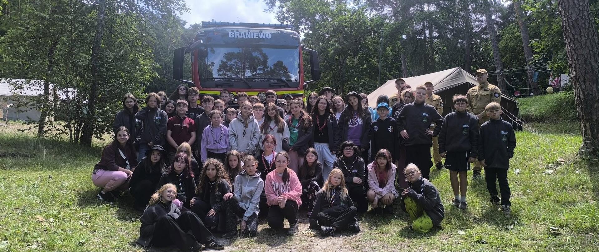 Na zdjęciu widać dużą grupę dzieci i nastolatków pozujących do wspólnej fotografii na leśnej polanie. Grupa liczy około 50 osób. Większość uczestników ma na sobie ciemne bluzy i chusty, co sugeruje, że mogą należeć do drużyny harcerskiej lub brać udział w zorganizowanym obozie.
W tle stoi czerwony samochód strażacki. Po prawej stronie widoczne są dwa namioty oraz dwie osoby w beżowych mundurach, które mogą pełnić rolę instruktorów lub przedstawicieli służb. Całość otacza wysoki sosnowy las, co nadaje zdjęciu typowo obozowy charakter.
Osoby są ustawione w kilku rzędach – część stoi, część klęczy lub siedzi z przodu.
Słonecznie.