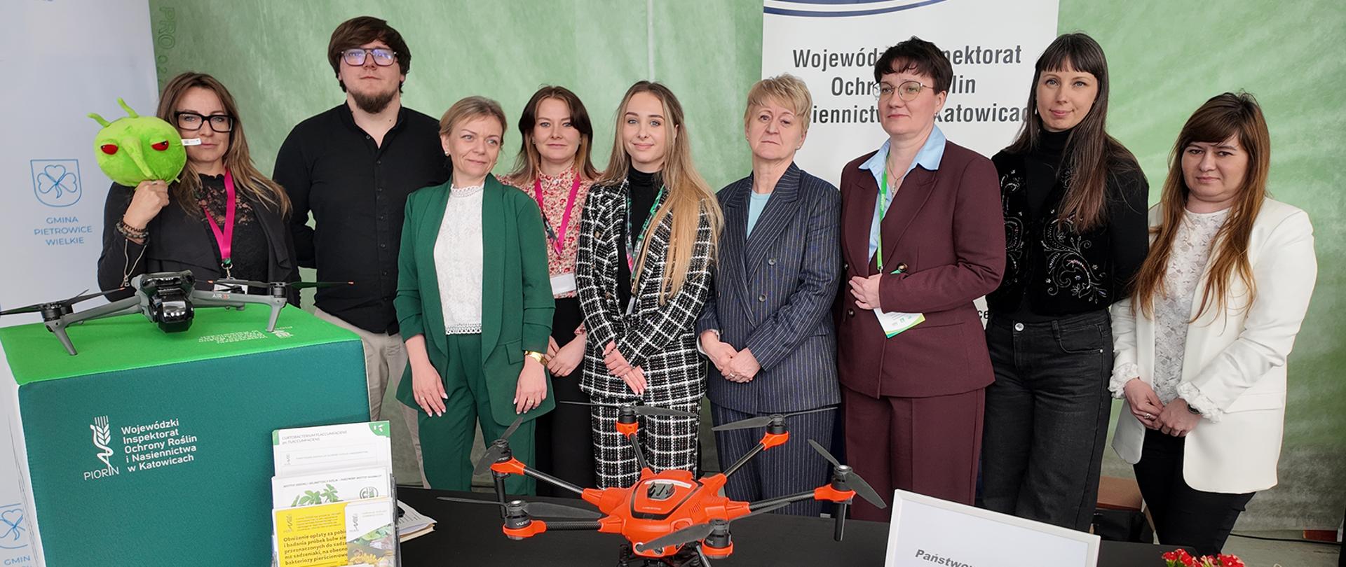 Reprezentanci WIORiN Katowice na stoisku informacyjnym. Zdjęcie przedstawia grupę osób 8 kobiet, 1 mężczyznę stojących w rzędzie, pozujących do zdjęcia. Na pierwszym planie widoczny jest dron.
