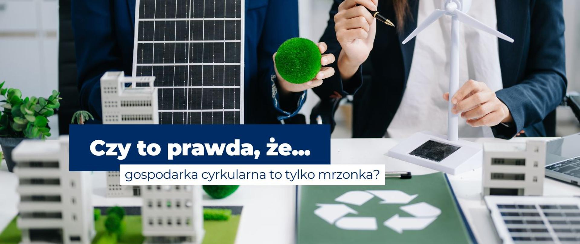 na zdjęciu widoczne są dłonie kobiet, które wykonują makietę OZE, na biurku leży kartka z symbolem recyklingu. Na środku baneru widnieje napis: Czy to prawda, że gospodarka cyrkularna to tylko mrzonka?