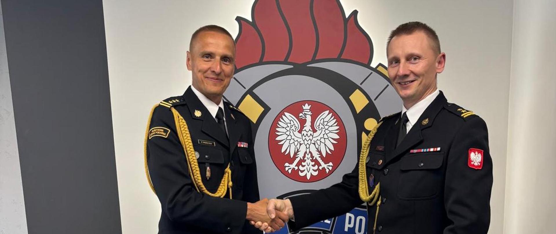 Na zdjęciu widzimy dwóch elegancko ubranych funkcjonariuszy Państwowej Straży Pożarnej, stojących na tle oficjalnego logotypu jednostki. Ujęcie przedstawia moment wręczenia dokumentu, co sugeruje ważne wydarzenie, takie jak awans, wyróżnienie lub uroczyste mianowanie.
