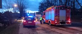 Widać dwa samochody straży pożarnej, na drodze rozsypany sorbent, w tle widać kilku strażaków oraz uszkodzony samochód