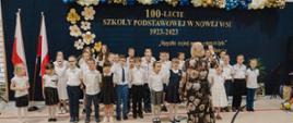W szkolnej sali gimnastycznej pod ścianą na której wisi czarny materiał z napisem 100-lecie Szkoły Podstawowej w Nowej Wsi 1923-2023 stoi grupa ubranych na galowo dzieci, przed nimi kobieta w sukience w kwiaty.