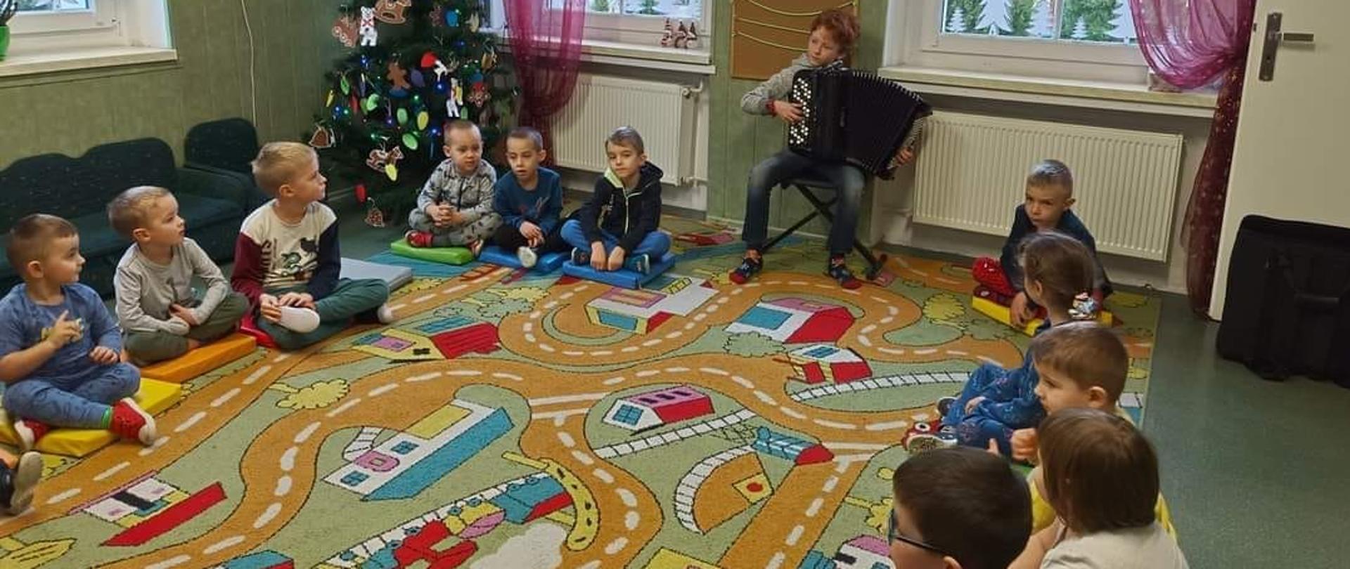 Uczeń klasy akordeonu Gabriel Charęza gra na akordeonie w przedszkolu w Mąkolnie. Wokoł niego siędzą mali chłopcy słuchający grę na instrumencie.