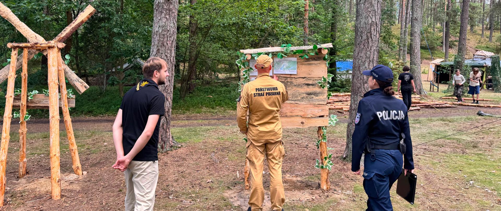 Na zdjęciu widoczni są: funkcjonariusz straży pożarnej, policjantka oraz kierownik obozu. Strażak sprawdza tablicę z informacjami o ewakuacji. W tle namioty