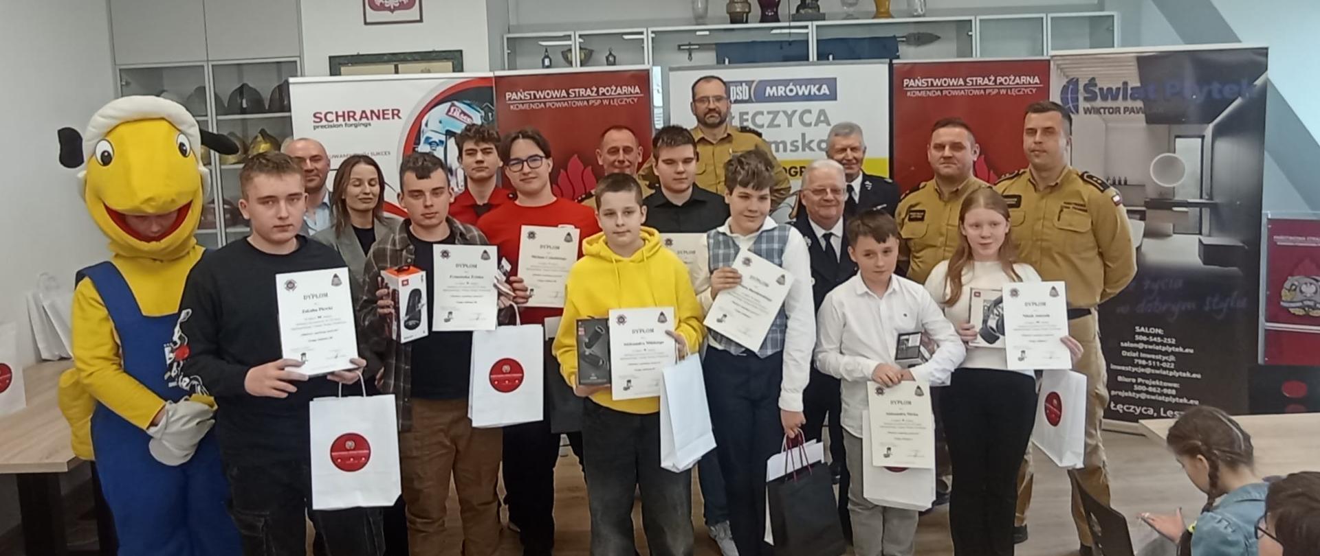 Na zdjęciu grupkę dzieci oraz dorosłych osób, którzy pozują do zdjęcia. Dzieci trzymają w rękach torebki oraz dyplomy. Wśród dorosłych jest jedna kobieta i siedmiu mężczyzn. Kobieta i jeden mężczyzna ubrani są w cywilne ubrania. Czterech mężczyzn ma założone piaskowy strój Państwowej Straży Pożarnej a dwóch ciemny ochotniczej straży pożarnej. Na zdjęciu widać jeszcze jedną osobę, która przebrana jest za żółtą mrówkę ubraną w niebieskie ubranie oraz dwójkę dzieci, które siedzą przy stole. W tle widać banery reklamowe i szafę z pucharami. Zdjęcie zrobione jest w pomieszczeniu, gdzie zapalone jest światło.