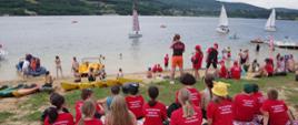 Widok z tyłu. Młodzieżowa drużyna pożarnicza siedzi na nadbrzeżu, niżej plażowicze. Na jeziorze żaglówki i pontony. 