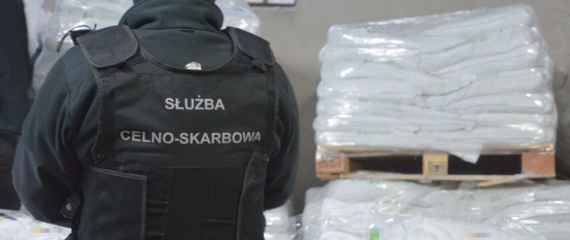 Funkcjonariusz Służby Celno-Skarbowej stojący tyłem, w tle zabezpieczone narkotyki.