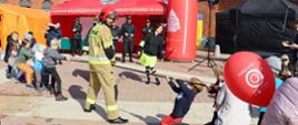 dzieci przeciągające linę, po środku przy linie stoi ubrany w ubranie bojowe i czerwony hełm strażak, w tle namiot straży pożarnej oraz stolik przykrytymi zielonym obrusem, obok trzech strażaków w ubraniu sztabowym przyglądają się konkurencji. W tle widoczne fragmenty budynków Manufaktury