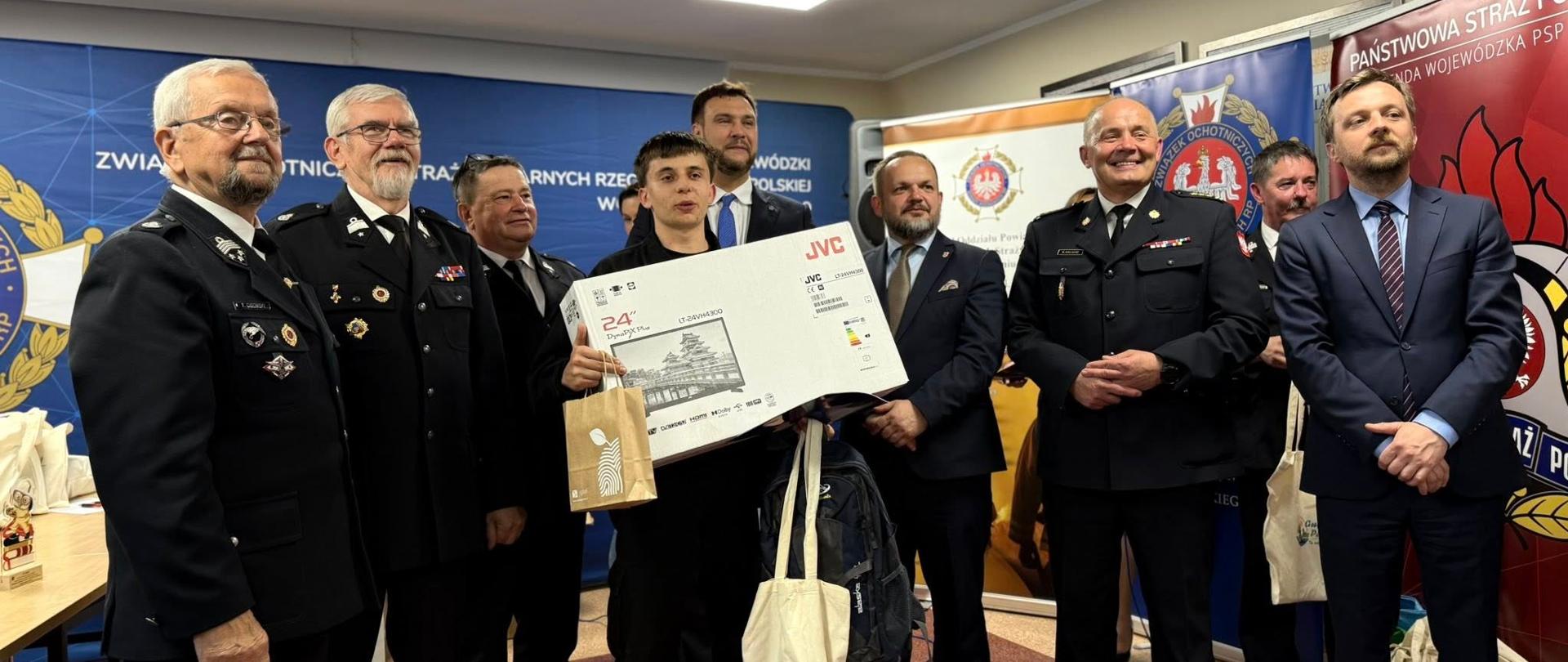 Grupa strażaków, przedstawicieli instytucji oraz laureat turnieju OTWP podczas wręczenia nagród. Uczestnik trzyma dużą nagrodę – monitor. W tle bannery Związku OSP RP i Komendy Wojewódzkiej PSP w Łodzi.