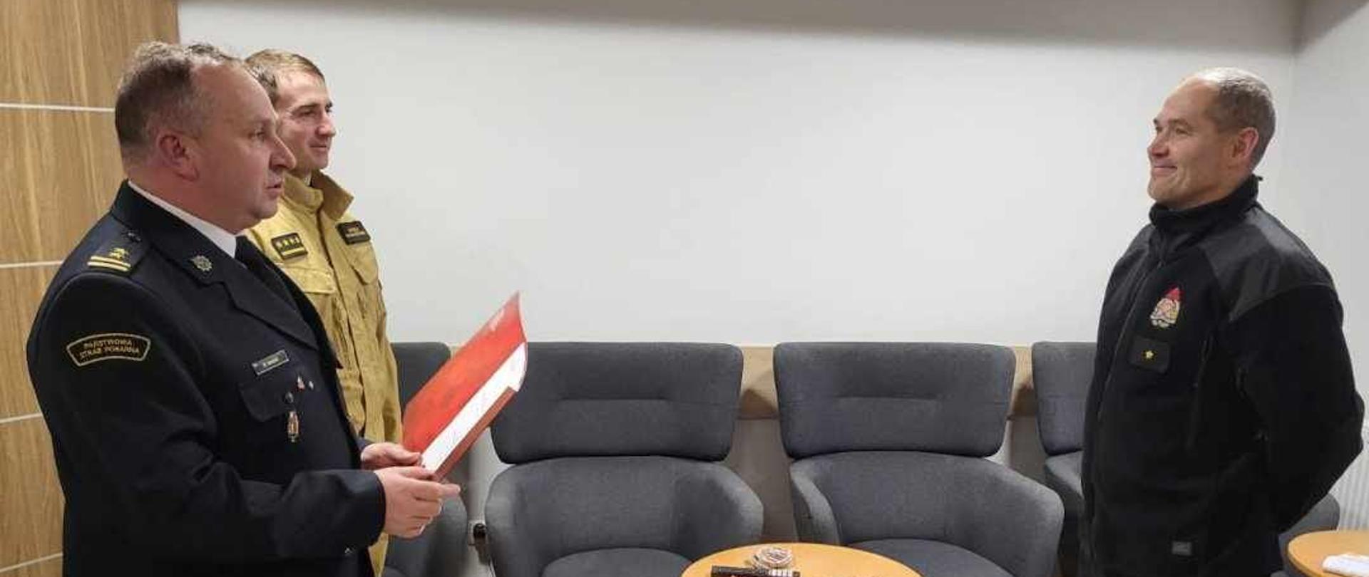 Komendant Powiatowy PSP w Pucku, bryg. Wojciech Wanat, odczytuje akt mianowania na stanowisko Zastępcy Dowódcy Zmiany dla asp. Mariusza Roeske. Komendant ubrany jest w mundur galowy wyjściowy bez złotego sznura, natomiast asp. Mariusz Roeske ma na sobie ubranie koszarowe. Na zdjęciu widoczny jest również Dowódca JRG w Pucku, st. kpt. Maciej Humanowski, ubrany w strój służbowy w kolorze piaskowym.