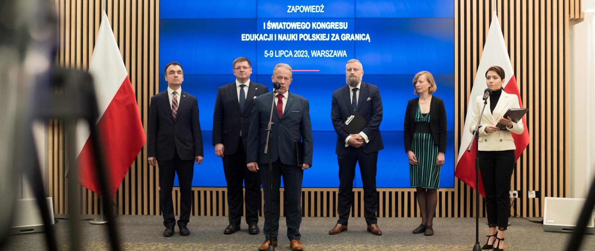 Na tle niebieskiej ściany - ekranu z napisem Zapowiedź I światowego kongresu edukacji i nauki polskiej za granicą stoi czterech mężczyzn w garniturach z których jeden mówi do mikrofonu na stojaku, i dwie kobiety - jedna w białej, druga w czarnej marynarce, po obu stronach ekranu polskie flagi.