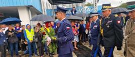 Policjant idąc trzyma wiązankę, za nim idą komendant wojewódzki Policji w Gdańsku pomorski komendant wojewódzki Państwowej Straży Pożarnej przedstawiciel wojska obok pod parasolami stoją zaproszeni goście.