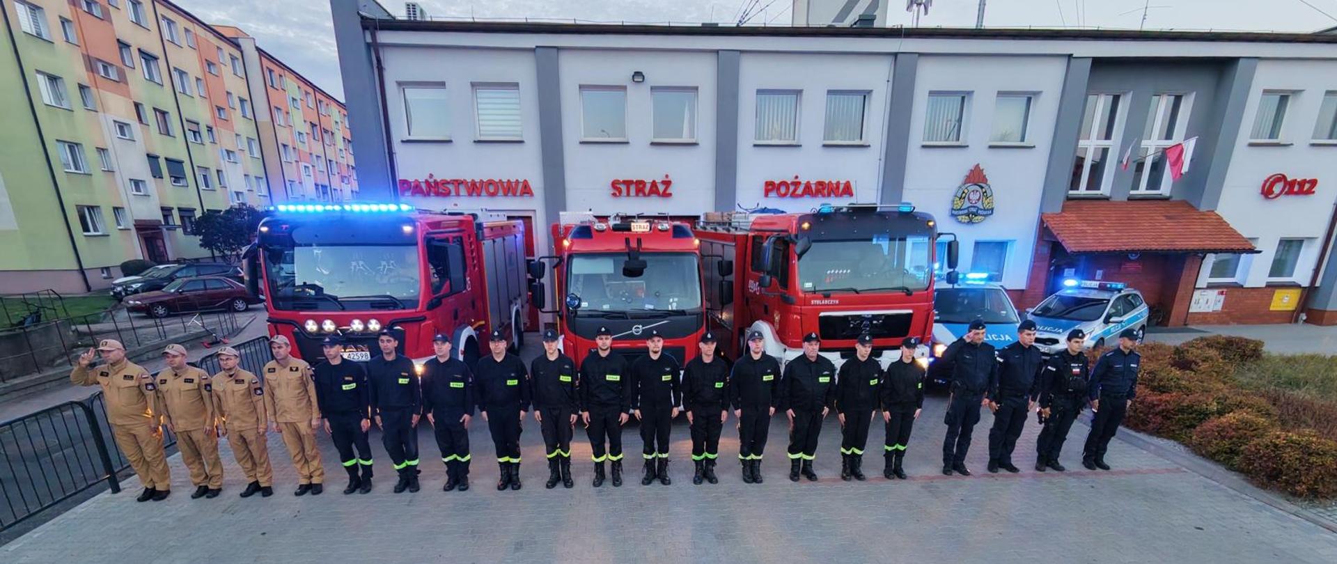 Strażacy i funkcjonariusze Policji pozują do wspólnego zdjęcia przed budynkiem Komendy Powiatowej Państwowej Straży Pożarnej w Krotoszynie. Za nimi stoją trzy czerwone samochody pożarnicze oraz radiowozy z włączonymi sygnałami świetlnymi. Funkcjonariusze oddają hołd zmarłym tragicznie kolegom. 