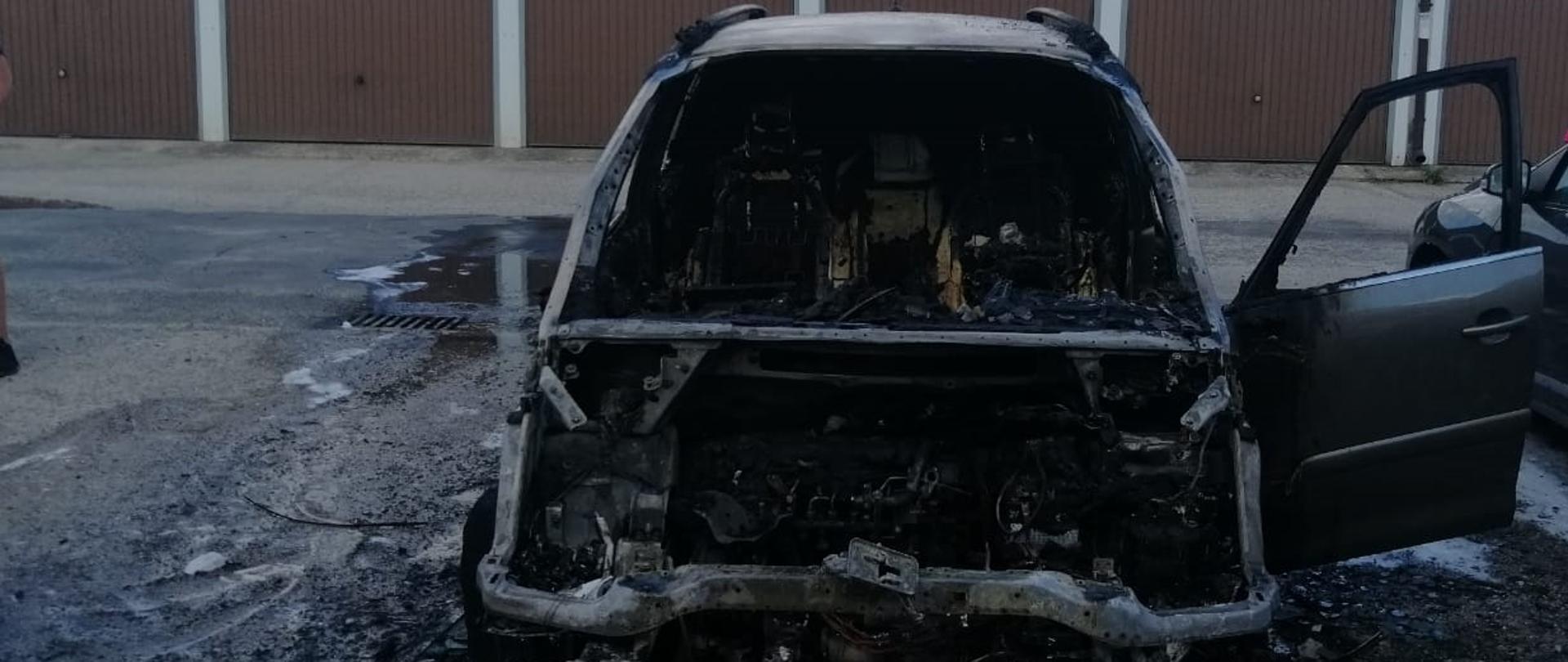 Widok z przodu na całkowicie spalą komorę silnika i wnętrze samochodu osobowego. W tle garaże.