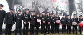 Zdjęcie przedstawia druhów z OSP Rąbino ubranych w mundury wyjściowe trzech strażaków trzyma plakaty z napisem "100-lecie niepodległości". Zdjęcie wykonano na ścianie, którą pomalowano artystyczne na 100-lecie niepodległości Rzeczypospolitej Polski.