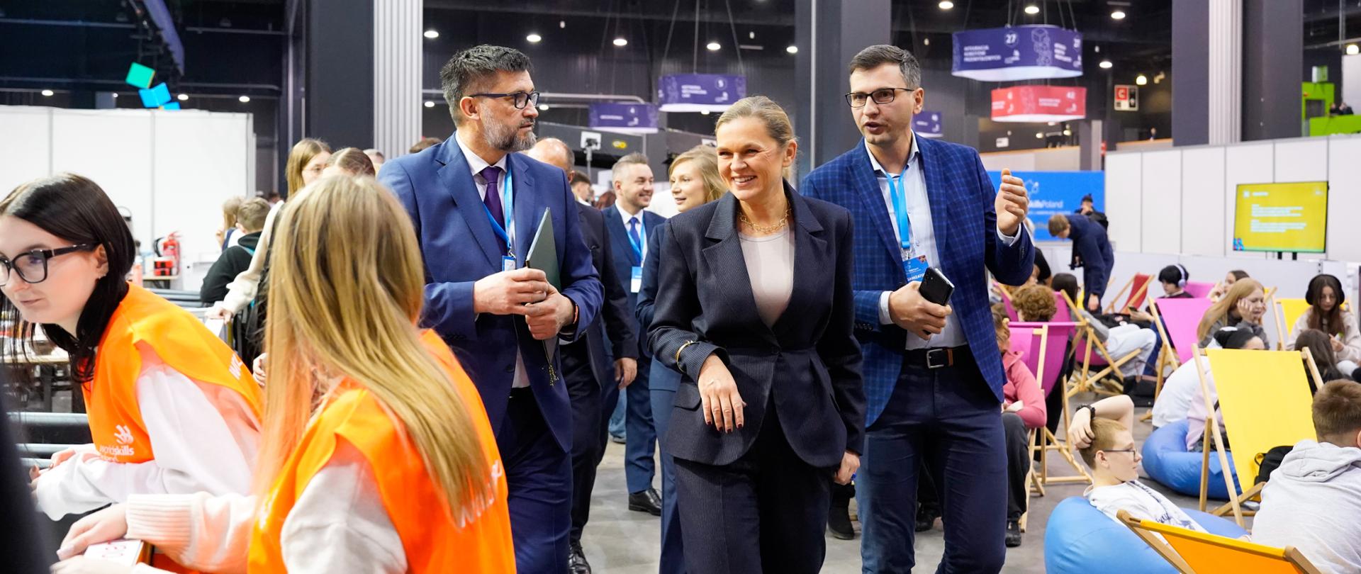 Kobieta w ciemnym garniturze (Barbara Nowacka) idzie przez otwartą halę wystawową w towarzystwie dwóch mężczyzn w garniturach. W tle widać innych uczestników i strefy relaksu z kolorowymi pufami.