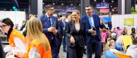 Kobieta w ciemnym garniturze (Barbara Nowacka) idzie przez otwartą halę wystawową w towarzystwie dwóch mężczyzn w garniturach. W tle widać innych uczestników i strefy relaksu z kolorowymi pufami.
