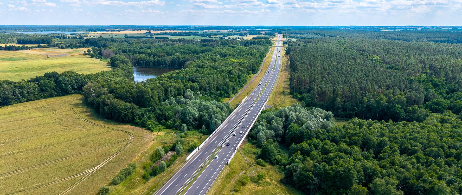 Widok z lotu ptaka na drogę ekspresową S3 przebiegającą przez rozległe tereny leśne i rolnicze. Kilka samochodów porusza się po dwujezdniowej trasie, która przecina zielone lasy, pola uprawne oraz niewielki zbiornik wodny. Zdjęcie wykonano w słoneczny dzień przy częściowym zachmurzeniu.
