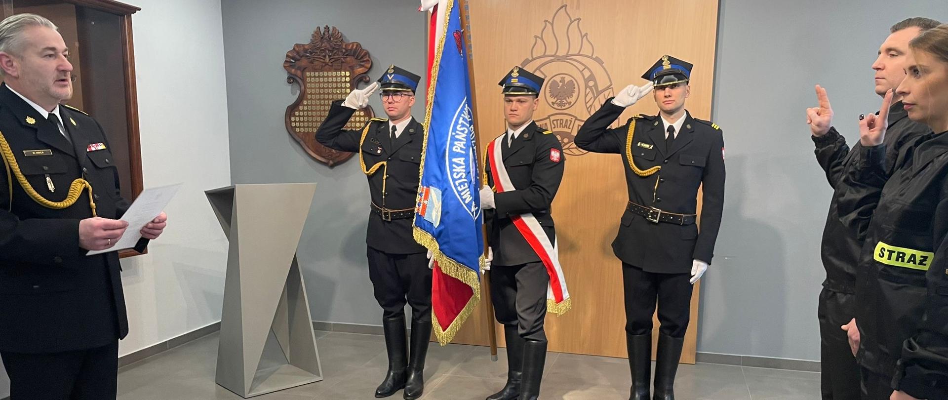 Świetlica komendy. Na wprost przy logo PPS stoi poczet sztandarowy. Po lewej komendant Po prawej nowi strażacy z podniesionymi prawymi dłońmi do ślubowania