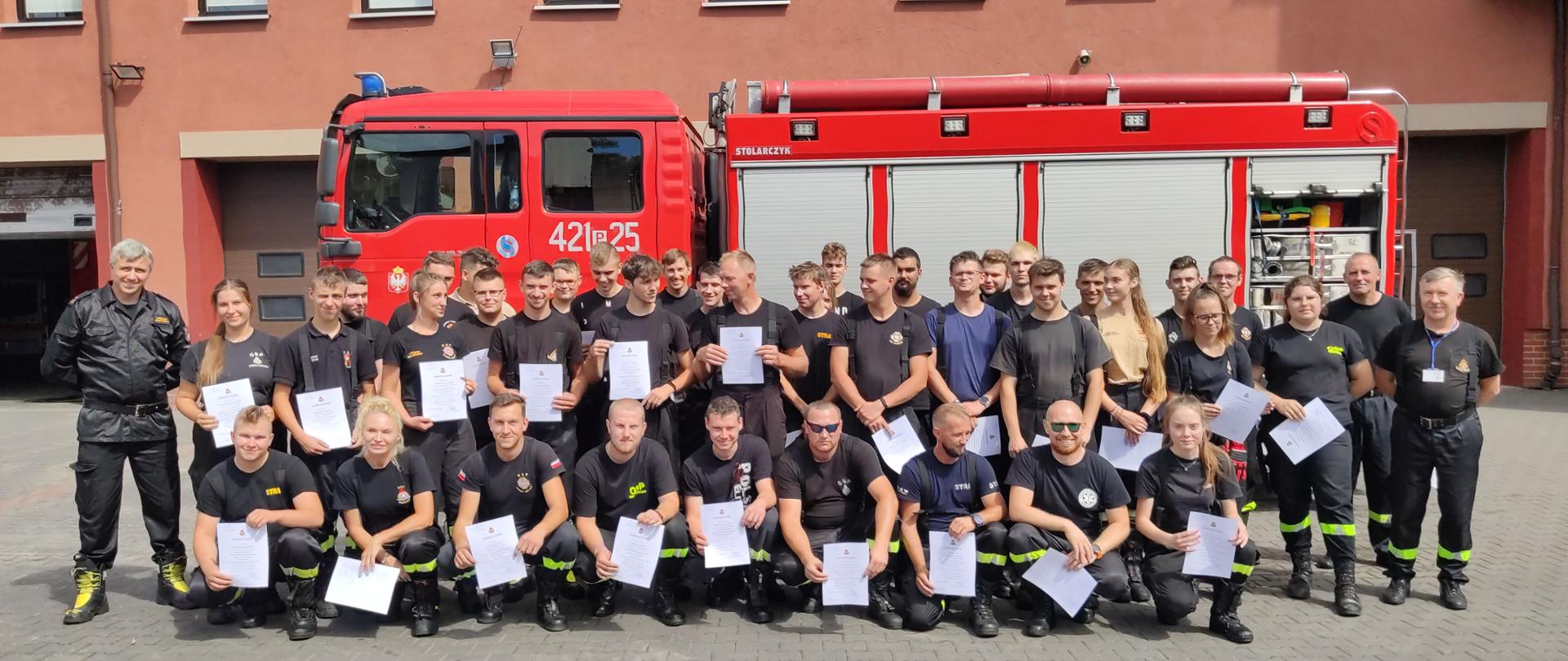 Zdjęcie grupowe wszystkich kursantów tuż po wręczeniu zaświadczeń o ukończeniu kursu. Zdjęcie zrobione jest na tle samochodu pożarniczego