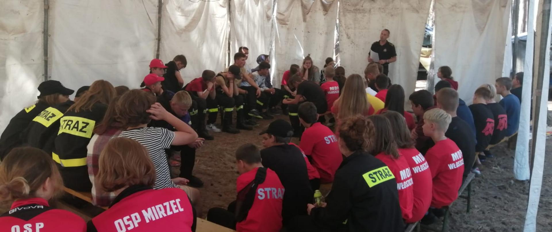 Zdjęcie przedstawia młodzież w namiocie na szkoleniu prowadzonym przez strażaka.