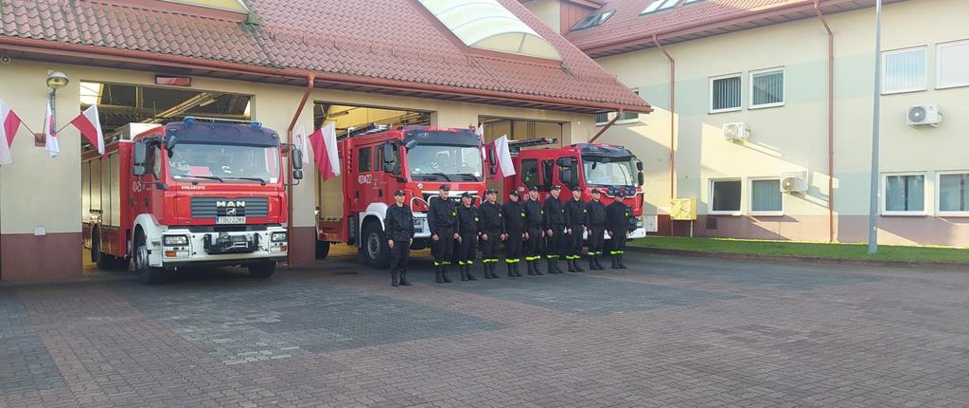 Uroczysta zbiórka strażaków przed budynkiem. W tle samochody pożarnicze.