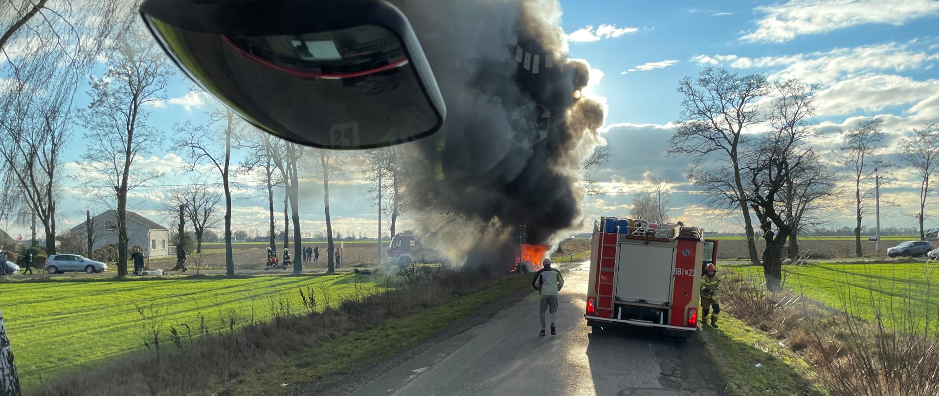 Zdjęcie przedstawia pożar dwóch samochodów osobowych po wypadku na drodze widoczny samochód strażacki jedna osoba postronna z boku pojazdu czarny dym oraz ogień z samochodów przy drzewach w oddali widać inny srebrny samochód oraz drzewa