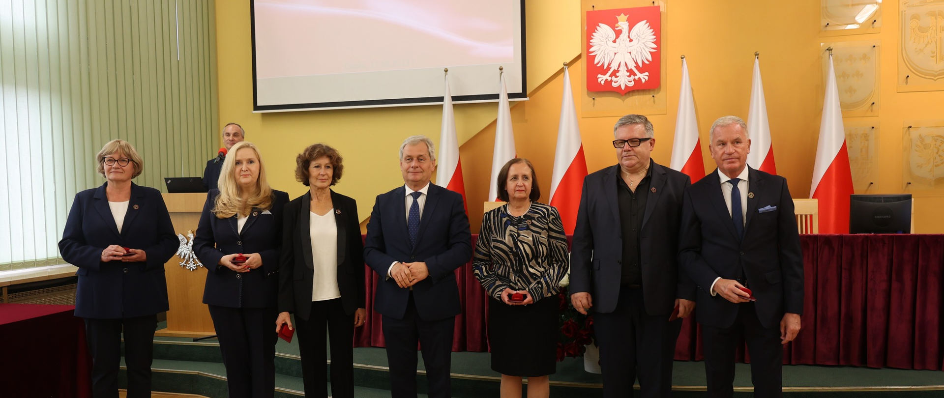 Zgromadzenie osób odznaczonych państwowymi odznaczeniami stojących. W tle biało-czerwone flagi polskie i godło polskie.
