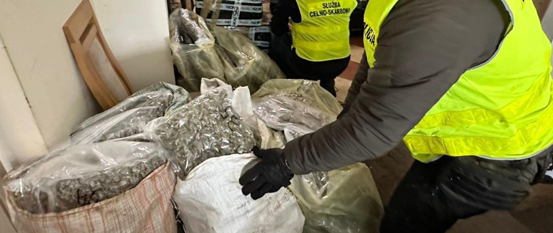 Osoby w kamizelkach służbowych stoją obok dużych worków wypełnionych suszem roślinnym.