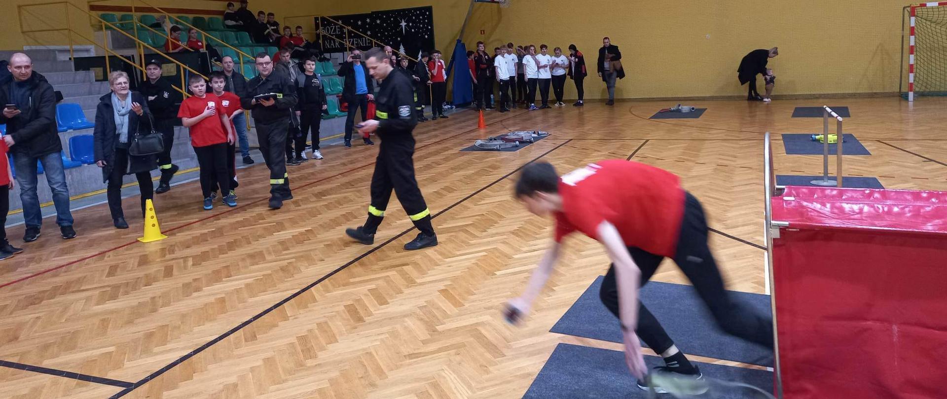Sala gimnastyczna na sali dzieci biorące udział w spartakiadzie obok funkcjonariusz na trybunach osoby postronne