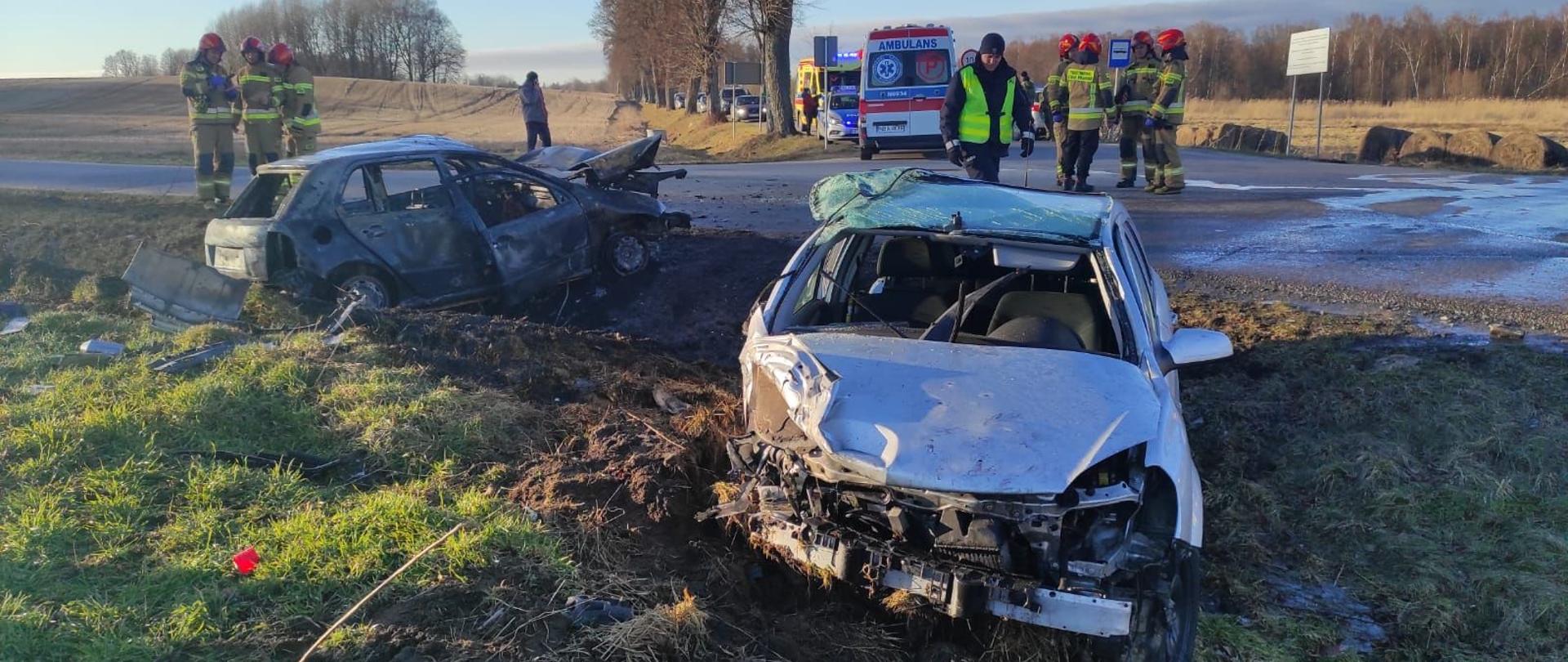 Na poboczu drogi dwa uszkodzone auta po zderzeniu, jeden z lewej cały spalony. Za nimi na drodze strażacy w mundurach i czerwonych hełmach. W oddali widać samochody karetki pogotowia. Słonecznie.