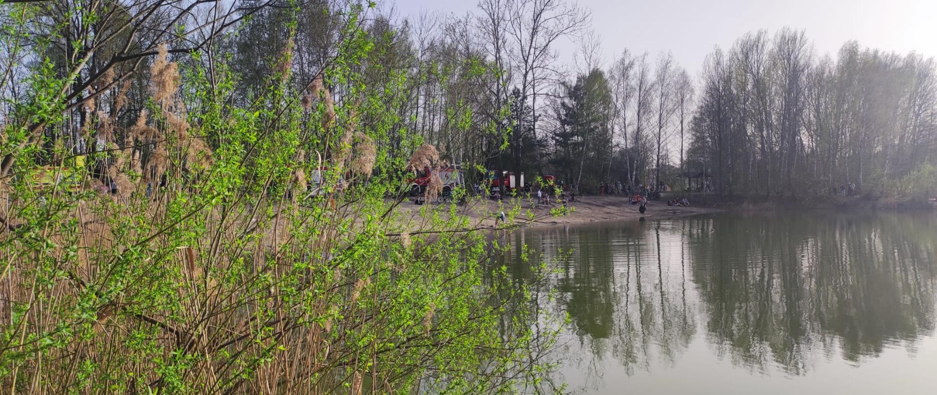 Na zdjęciu widoczna jest zbiornik wodny a w oddali ustawione pojazdy służb ratunkowych częściowo zasłonięte przez krzewy.